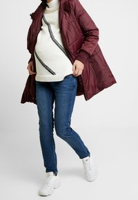 Maroon padded coat with a high collar, worn over a white knit sweater featuring black diagonal stripes, paired with blue jeans and white sneakers.