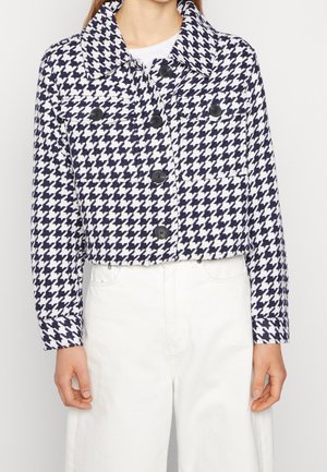 Femme portant une veste courte à motif pied-de-poule bleu marine et blanc avec des boutons noirs, associée à un pantalon blanc taille haute.