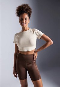 Jovem mulher com cabelo encaracolado usa uma t-shirt cropped bege e calções de ciclista castanhos de cintura alta, de pé com a mão na anca contra um fundo cinza.