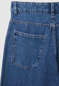 Jean en denim bleu moyen avec une poche arrière de forme rectangulaire. Le tissu présente un motif tissé texturé et des coutures visibles.