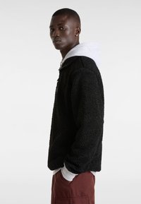 Chaqueta de forro polar negro con textura y capucha, superpuesta a una sudadera gris. Presenta un bolsillo frontal y un ajuste relajado.