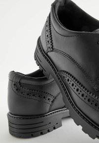 Chaussures en cuir noir avec des perforations décoratives, bout arrondi, semelles à relief robustes et surpiqûres contrastantes le long des bords.