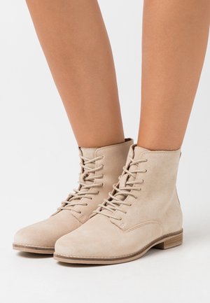 Bottes cheville en daim beige avec un bout arrondi, un laçage à l'avant et un talon bas empilé. Texture lisse et détails de couture minimalistes.