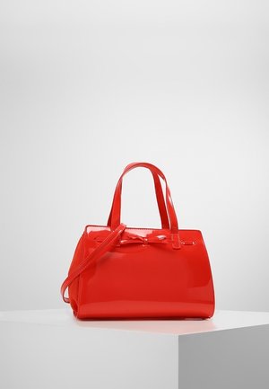 Sac à main rouge brillant avec deux poignées supérieures, une longue bandoulière, et un petit nœud décoratif sur le devant, présenté sur une plateforme blanche.