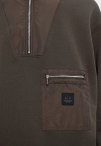 Pullover verde oliva con zip a V, dotato di una tasca con zip argentata e etichetta del logo. Tessuto morbido con una superficie liscia.