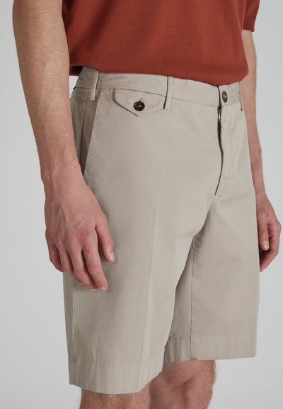Uomo che indossa pantaloni corti beige su misura con tasca a patta con bottone e una camicia color ruggine, in piedi di fronte a uno sfondo neutro.