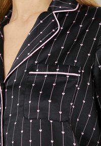 Zwart satijnen pyjamapakket met roze hartpatronen en verticale lijnen. Voorzien van een notch-kraag en een borstzak, omlijnd met roze bies.