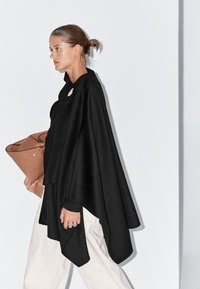 Žena s hladce učesanými vlasy, oblečená v černém plášti a bílých kalhotách, držící velkou hnědou koženou tašku, kráčející proti bílé zdi.