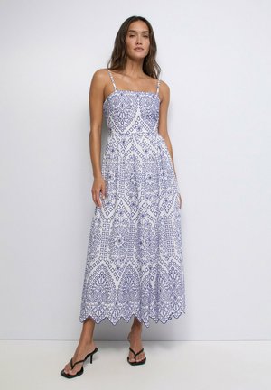 Mujer de pie contra una pared, vestida con un vestido largo blanco sin mangas con patrones geométricos y florales azules y sandalias negras de tacón.