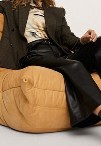 Vaaleanruskea ylisuuri pussituoli, jossa on pehmeä pinta ja pyöreä muoto, ja sen päällä istuu henkilö, jolla on tumman ruudullinen bleiseri ja mustat nahkahousut.