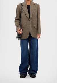 Blazer a spina di pesce marrone, jeans in denim blu larghi, top nero e stivaletti con suola spessa. Piccola borsa nera con frange.