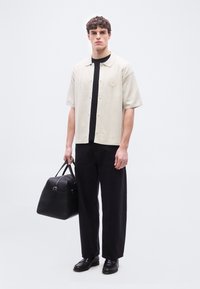 Νεαρός άνδρας που στέκεται, φοράει ανοιχτό μπεζ κοντομάνικο πουκάμισο πάνω από μαύρο μπλουζάκι, μαύρο παντελόνι, μαύρα παπούτσια, κρατώντας μαύρη δερμάτινη τσάντα duffel στο δεξί χέρι.