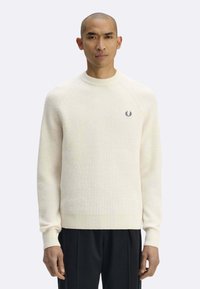 Maglione color crema a costine con scollo rotondo, presenta un piccolo logo blu sul petto a sinistra. Indossato con pantaloni neri.