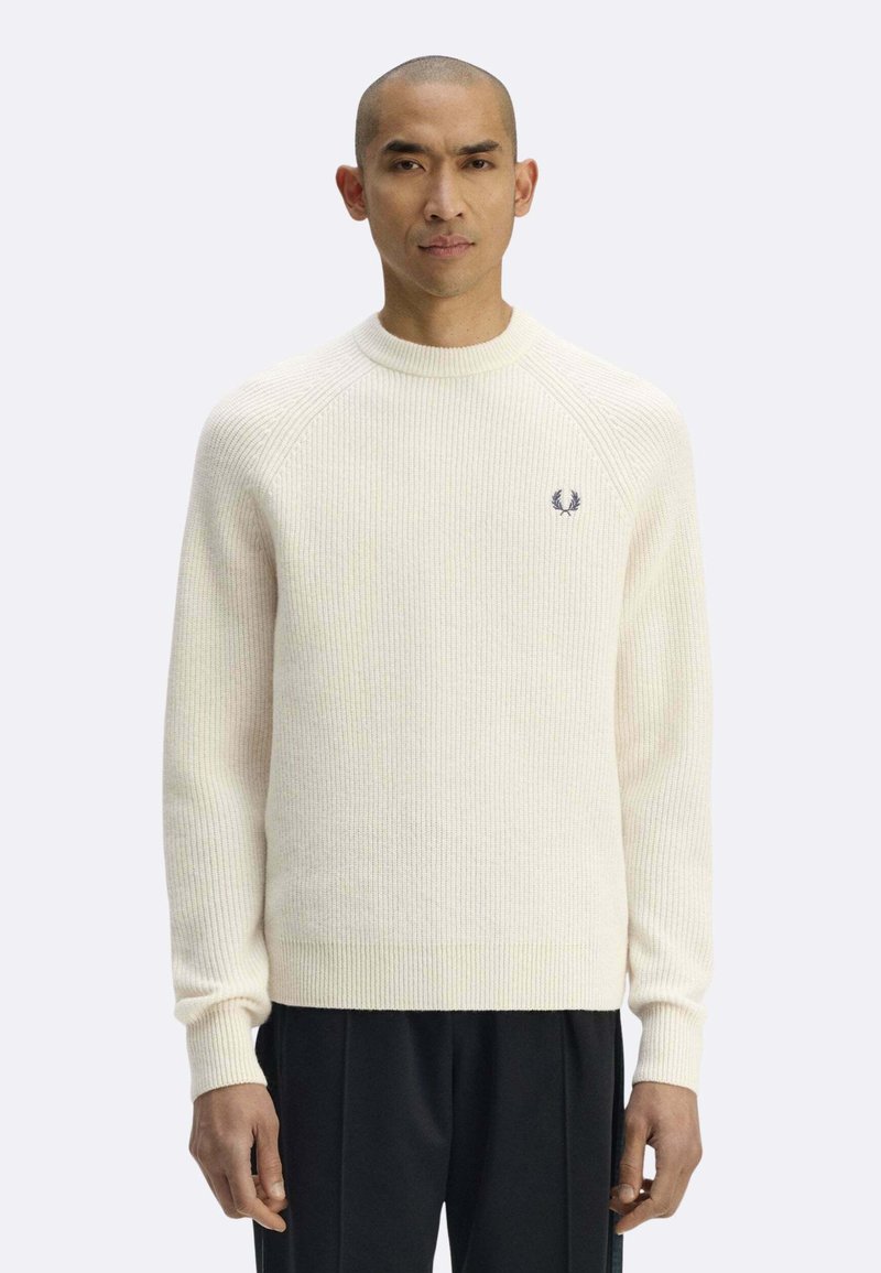 Maglione color crema a costine con scollo rotondo, presenta un piccolo logo blu sul petto a sinistra. Indossato con pantaloni neri.