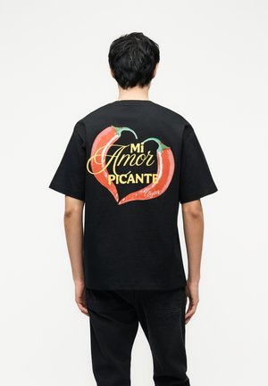 Mężczyzna w czarnej koszulce z dwoma czerwonymi papryczkami chili tworzącymi serce oraz żółtym napisem "Mi Amor Picante" na plecach.