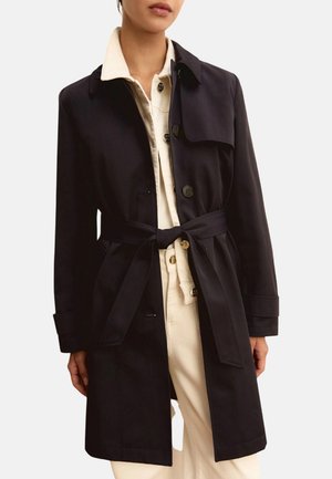 Donna che indossa un trench nero con cintura sopra una camicia crema con bottoni e pantaloni crema, in piedi davanti a uno sfondo bianco uniforme.