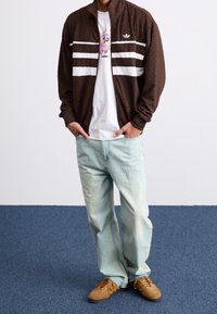 Ruskea vetoketjullinen takki, jossa on valkoisia raitoja, valkoisen t-paidan päällä, jossa on värikäs grafiikka. Vaaleansiniset löysät farkut ja ruskeat kengät.