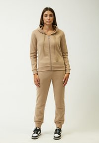 Felpa con zip beige, dotata di lacci, abbinata a pantaloni felpati a gamba affusolata dello stesso colore. Tessuto morbido, polsini a costine e sneakers nere con dettagli bianchi.