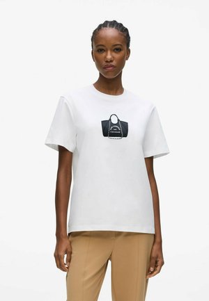 Woman wearing a white t-shirt with a black handbag graphic and beige pants, standing against a plain light background.