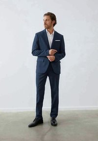 Fato azul marinho feito de tecido texturizado, com lapela de pico, botão único e lenço de bolso branco; combinado com uma camisa branca nítida e sapatos pretos.