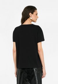 Zwarte T-shirt met korte mouwen en een relaxte pasvorm. Het materiaal lijkt zacht. De model draagt ook hartvormige gouden oorbellen, wat een subtiele accent toevoegt.