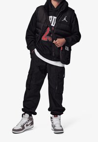 Gilet imbottito nero indossato sopra una felpa nera con grafica rossa "23", abbinato a pantaloni cargo neri e sneakers alte grigie. Borsa a tracolla nera.