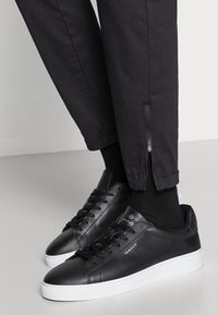 Zapatillas de cuero negro con suelas de goma blanca, que cuentan con una textura suave, diseño de cordones y detalle de logo. Pantalones negros con puños con cremallera.