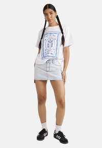 Witte katoenen t-shirt met blauw grafisch ontwerp, gecombineerd met een lichte denimrok. Zwarte sportschoenen met witte strepen en witte sokken.