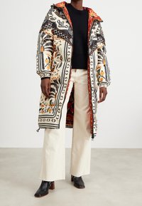 Abrigo acolchado con patrones florales y geométricos vibrantes en naranja, negro y crema. Cuenta con un forro de capucha rojo y dobladillo ajustable.