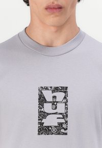 Grå kortermet T-skjorte med et svart grafisk logo omkranset av intrikate mønstre. Stoffet ser mykt og glatt ut.