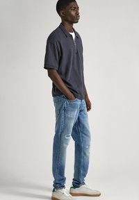 Polo de navy con cremallera, jeans claros desgastados y zapatillas blancas con detalles en verde. El modelo está de pie frente a un fondo neutral.