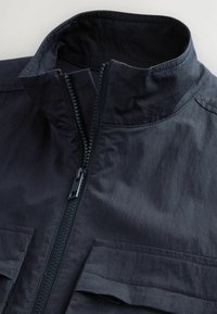 Giacca blu navy con colletto alto, chiusura con zip visibile e due tasche frontali con alette piatte, realizzata in tessuto morbido.