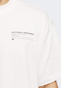 Camiseta de algodón blanca con una pequeña etiqueta negra impresa que dice "Anthony Edwards" y detalles de ingeniería en el área del pecho izquierdo.