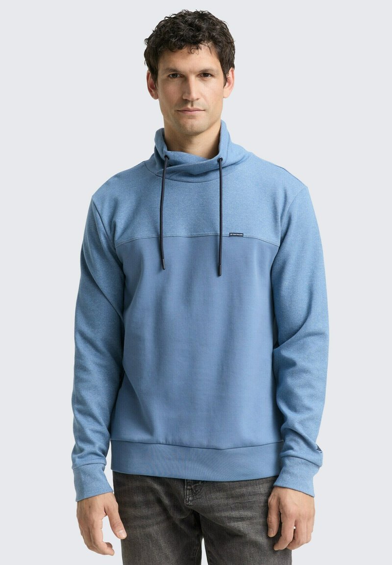 Sweat-shirt à col montant bleu clair avec cordon de serrage et panneau contrastant bleu plus foncé sur la poitrine. Texture de tissu douce.