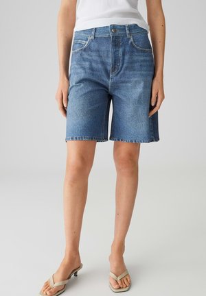 Vrouw draagt blauwe denim shorts tot op de knie, een wit tanktop en beige sandalen met hak, staand tegen een eenvoudige grijze achtergrond.