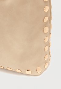 Sac en similicuir beige aux coins arrondis, orné d'étincelants clous métalliques le long d'un côté, offrant une texture lisse.
