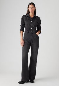 Juodas džinsinis kombinezonas su prigludusia liemene, ilgomis rankovėmis ir plačiomis flaro kelnėmis. Su priekinių kišenių ir mygtukų užsegimu. Lygi tekstūra.
