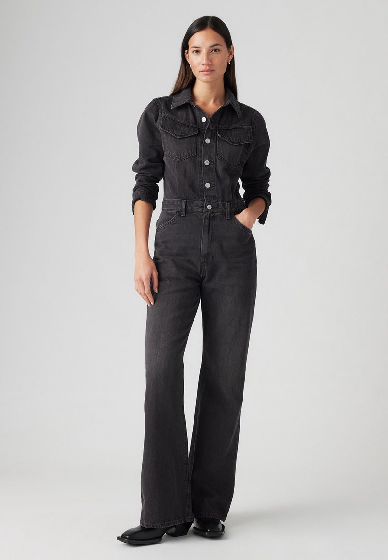 Juodas džinsinis kombinezonas su prigludusia liemene, ilgomis rankovėmis ir plačiomis flaro kelnėmis. Su priekinių kišenių ir mygtukų užsegimu. Lygi tekstūra.