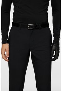 Skräddarskjuten långärmad svart skjorta, svart bälte med silverspänne och svarta byxor. Vänster hand har på sig en svart läderhandske. Enkel texturerad väv.