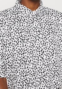 Chemise à manches courtes en tissu blanc avec un motif à taches noires, design avec col et fermeture à boutons.