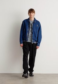 Veste en denim avec fermeture éclair, t-shirt graphique gris en dessous, pantalon cargo noir et chaussures noires, sur fond uni.