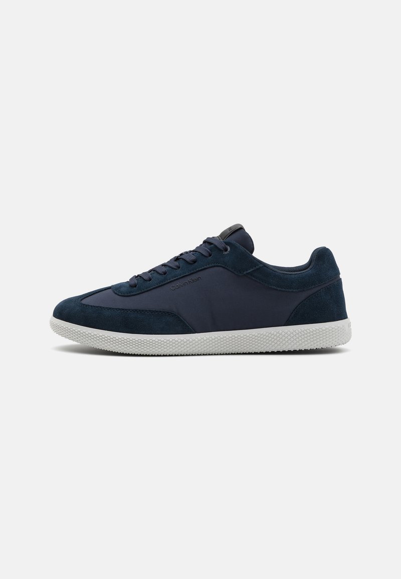 Mørkeblå sneakers laget av semsket skinn og stoff med en strukturert hvit gummisåle. Har snørebånd og en logo på siden, designet for hverdagsbruk.