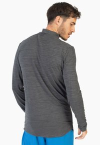 Camisa de manga larga en gris oscuro, confeccionada con un tejido texturizado, con un cuello alto y mínimos detalles de costura, llevada con pantalones azules.