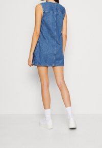 Kobieta stoi w niebieskiej, bezrękawnikowej mini sukience dżinsowej, z białymi skarpetkami i białymi trampkami na tle gładkiego, jasnego tła.