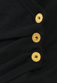 Peça de roupa de malha preta com três grandes botões dourados com texto em relevo. O tecido tem uma textura suave e um caimento sutil.