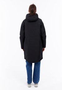 Sort hættetrøje med lang kant, glat tekstur og justerbar hætte, parret med blå jeans og lyse sko.