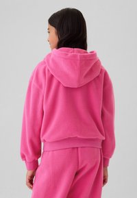 Mlada oseba, obrnjena stran, oblečena v svetlo roza flis pulover s kapuco in ujemajoče se hlače, na enostavni svetli podlagi.