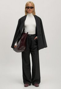 Vrouw in een zwarte oversized blazer en broek, witte turtleneck, kastanjebruine gestikte schoenen, zwarte zonnebril, en met een grote kastanjebruine tas.