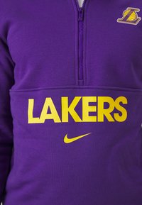 Pull-over violet avec un zip quart, "LAKERS" en lettres jaunes en gras, et un logo Nike en jaune ; texture de tissu lisse.