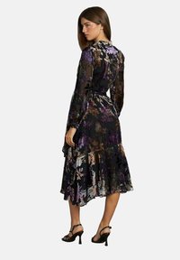 Vestido negro con flores, de mangas largas, con un dobladillo rizado y una cintura ajustada. Presenta patrones florales en púrpura, oro y blanco sobre una tela transparente.